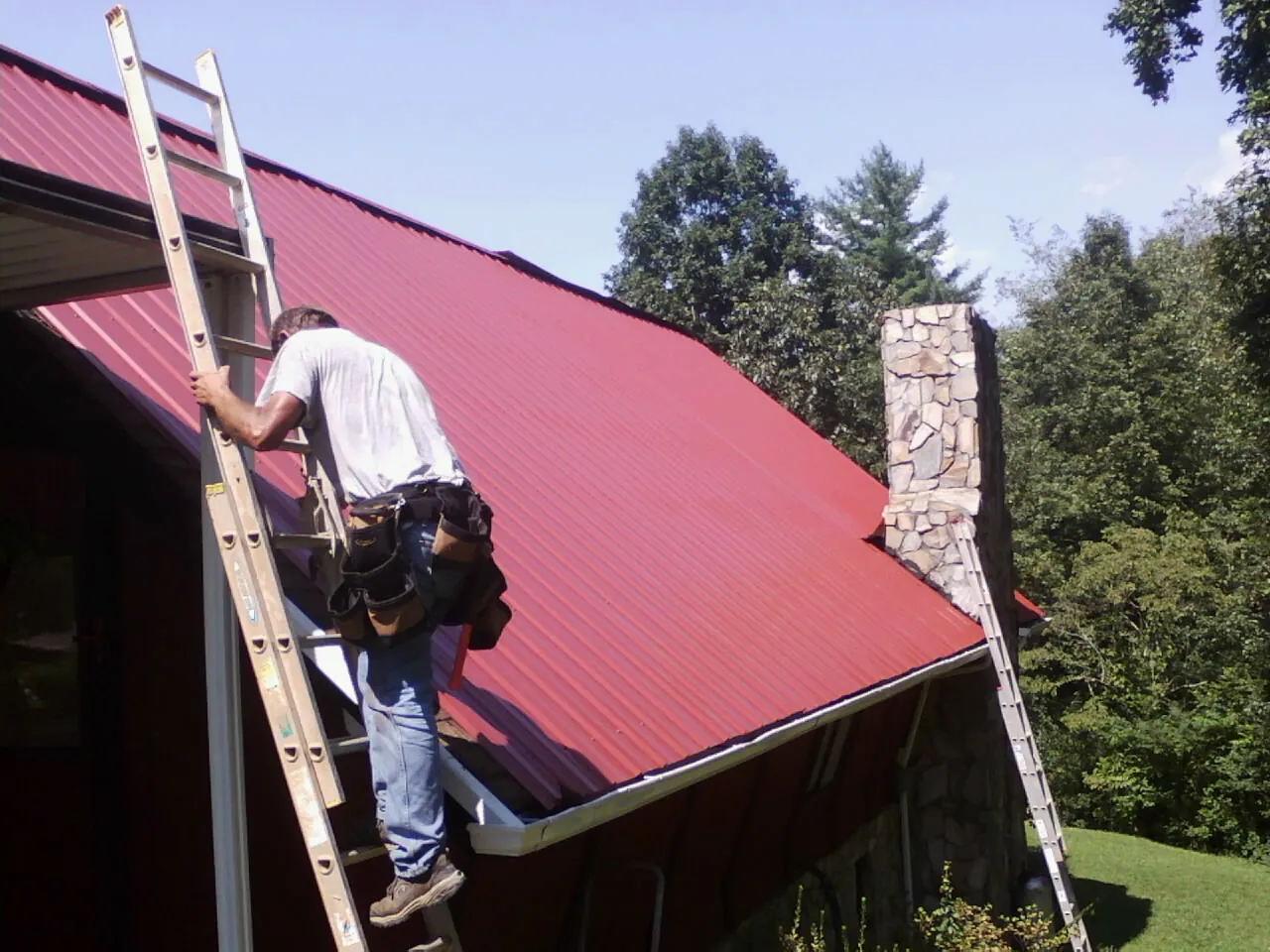 Roofer on a ladder beside a red metal roof and stone chimney — Soffit & Fascia Repair by Roofing Brothers in Northboro